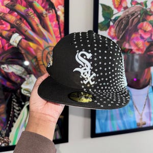 Pode incluir: Um bon&eacute; de beisebol preto com um logotipo 'S' branco e strass. O bon&eacute; tem uma aba preta e uma aba interna branca.