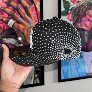 Pode incluir: Um bon&eacute; de beisebol preto com um logotipo branco e uma cobertura completa de strass transparentes.