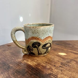 Puede incluir: Una taza de cerámica hecha a mano con un esmalte verde claro y detalles marrones. La taza presenta un diseño de setas en negro y marrón.