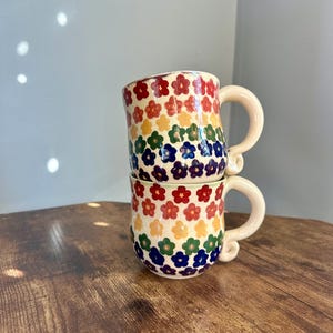 May include: Two ceramic mugs stacked on top of each other. Each mug is decorated with a pattern of colorful flowers in red, orange, yellow, green, blue, and purple. The mugs have cream-colored handles.