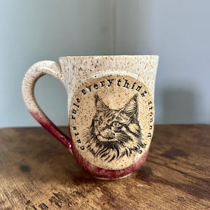 Peut inclure: Une tasse en céramique tachetée avec une base et une anse bordeaux. La tasse présente un motif circulaire avec une illustration détaillée de chat et le texte "cats rule everything around me."