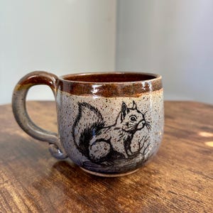May include: A ceramic mug with a brown handle and rim, featuring a black and white illustration of a squirrel. The mug has a speckled, light blue body and sits on a wooden surface. The mug is designed for hot beverages.