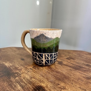 May include: A ceramic mug with a mountain landscape design. The mug features a speckled beige rim, a mountain scene in shades of green and gray, and a geometric pattern in navy blue. The handle is beige.