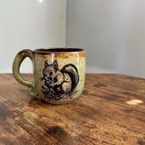 May include: A ceramic mug with a light green speckled glaze and a brown rim. The mug features a black and white illustration of a squirrel holding a nut. The handle is a light green color.