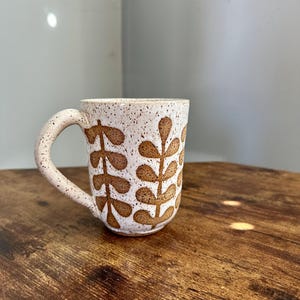 May include: A speckled ceramic mug with a brown leaf pattern. The mug has a curved handle and is set on a wooden surface. The mug's exterior is white with brown speckles and brown leaf designs.