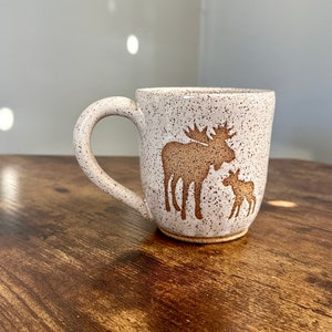 May include: A speckled ceramic mug with a brown moose and calf design. The mug has a curved handle and a speckled white and brown exterior. The moose and calf are in a brown silhouette. The mug is on a wooden surface.