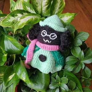 May include: A green and black crocheted plush toy with a pink scarf and glasses. The toy is sitting in a pot of green plants.