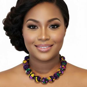 May include: A woman wearing a colorful beaded necklace. The necklace features large, round beads in shades of purple, yellow, green, and black. The woman has dark hair and is smiling. The necklace is a statement piece.