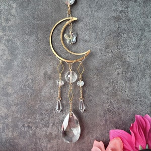 May include: A gold crescent moon-shaped metal ornament with a star and crystal beads hanging from it. The ornament has a crystal teardrop hanging from the bottom.
