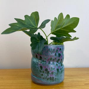 May include: A ceramic planter with a textured, mottled glaze in shades of teal, purple, and green. The cylindrical pot holds several large, green, leafy plants. The planter sits on a wooden surface.