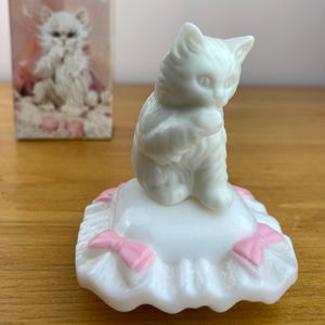 May include: A white ceramic kitten figurine sitting on a white pillow with pink bows. The kitten is in a seated position with its paws together. A box with a picture of the kitten is in the background.