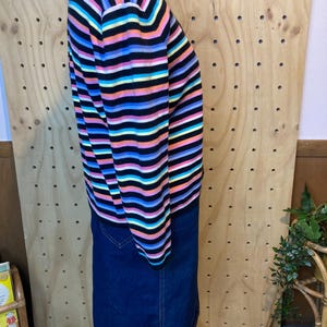 May include: A long-sleeved sweater with horizontal stripes in black, pink, orange, yellow, blue, and purple. The sweater is paired with a dark blue denim skirt. The clothing is displayed on a mannequin.
