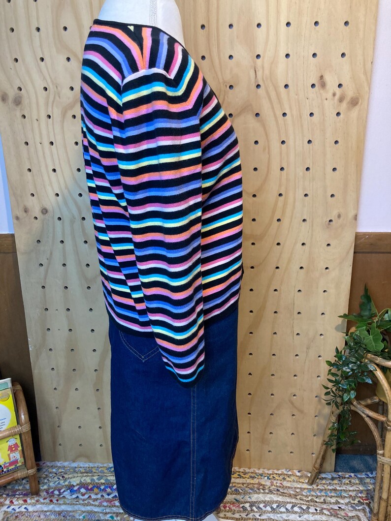 May include: A long-sleeved sweater with horizontal stripes in black, pink, orange, yellow, blue, and purple. The sweater is paired with a dark blue denim skirt. The clothing is displayed on a mannequin.