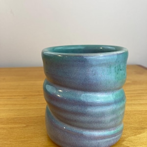 May include: A handmade ceramic tumbler with a unique, swirled design. The cup features a gradient of teal and purple hues, with a glossy finish. The pottery is set on a wooden surface.