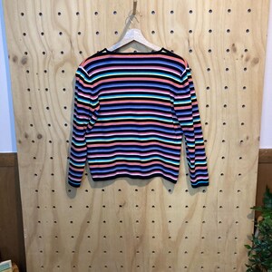 May include: A long-sleeved sweater with horizontal stripes in various colours, including black, pink, orange, blue, and teal. The sweater is hanging on a wooden hanger against a light wood background.