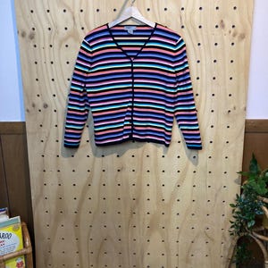 May include: A colourful striped cardigan with a V-neck and button fastening. The sweater has horizontal stripes in black, pink, blue, and teal. The long-sleeved cardigan is hanging on a wooden hanger against a wooden pegboard.
