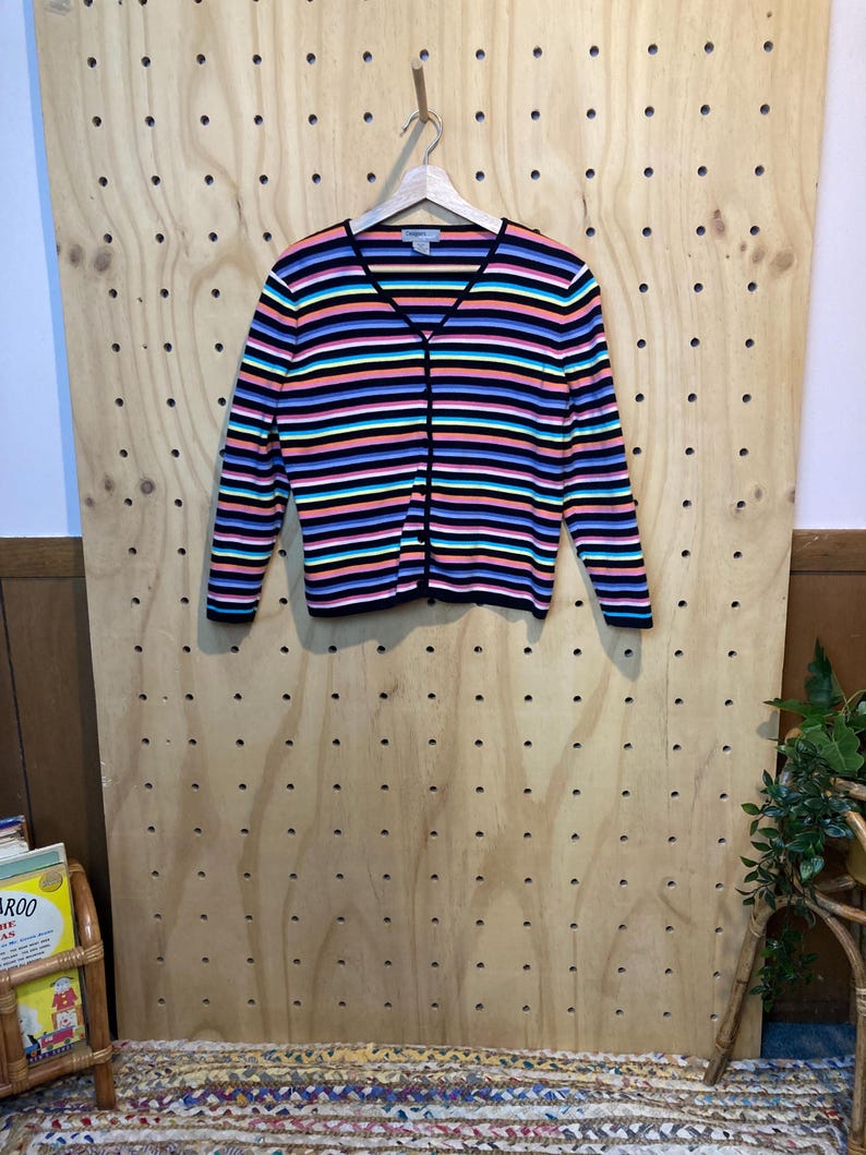 May include: A colourful striped cardigan with a V-neck and button fastening. The sweater has horizontal stripes in black, pink, blue, and teal. The long-sleeved cardigan is hanging on a wooden hanger against a wooden pegboard.