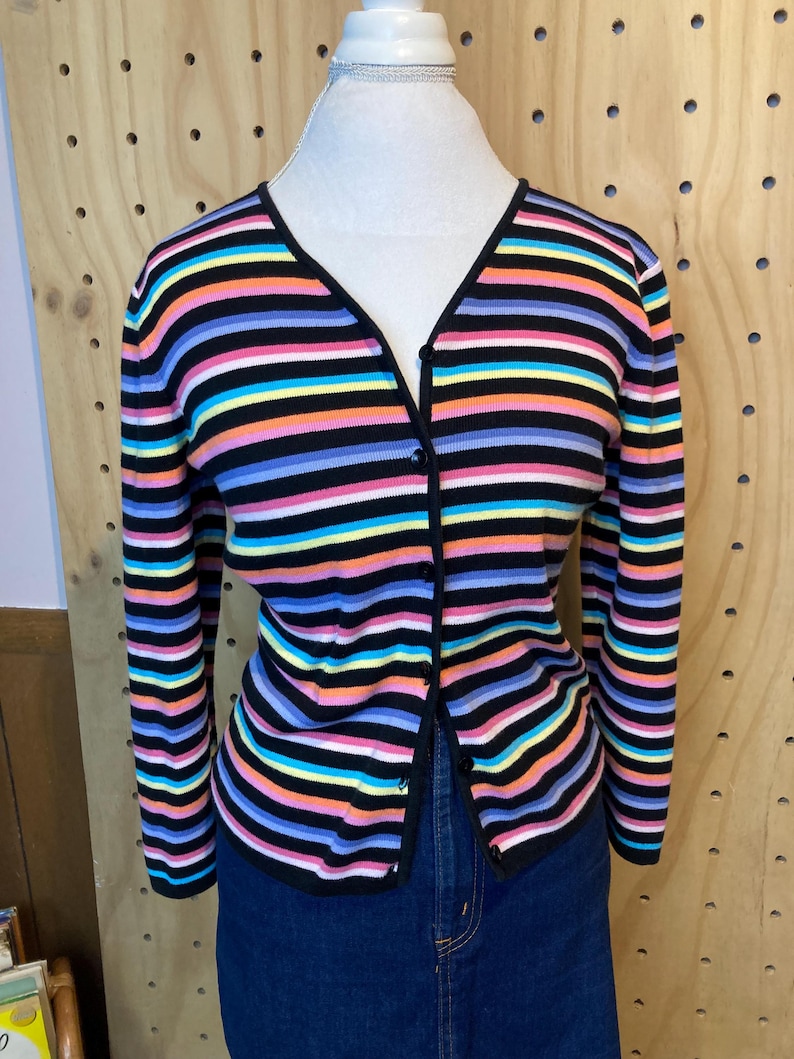 May include: A colourful striped cardigan with a V-neck and long sleeves. The sweater has horizontal stripes in black, pink, orange, yellow, blue, and purple. The cardigan is buttoned up and displayed on a mannequin.