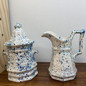 May include: A white ceramic sugar bowl with a lid and a matching pitcher. Both pieces are decorated with blue and black splatter paint. The sugar bowl has two small handles, and the pitcher has a handle and a spout.