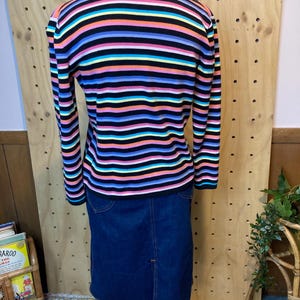 May include: A multi-coloured striped sweater with long sleeves, in shades of black, pink, blue, yellow, and green, is worn with a dark blue denim skirt. The sweater has horizontal stripes. The skirt has a split at the back.