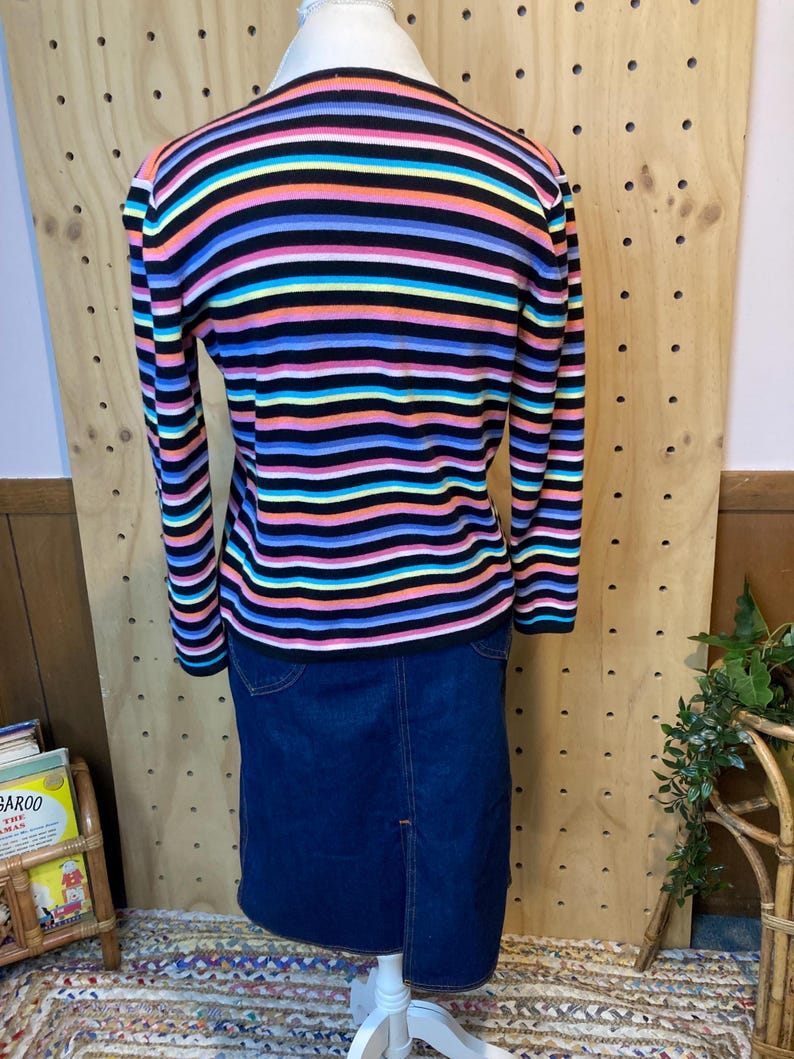 May include: A multi-coloured striped sweater with long sleeves, in shades of black, pink, blue, yellow, and green, is worn with a dark blue denim skirt. The sweater has horizontal stripes. The skirt has a split at the back.