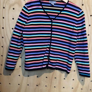 May include: A colourful striped cardigan with a V-neck and long sleeves. The sweater has horizontal stripes in black, pink, blue, yellow, orange, and white. The cardigan is buttoned up and hanging on a wooden hanger.