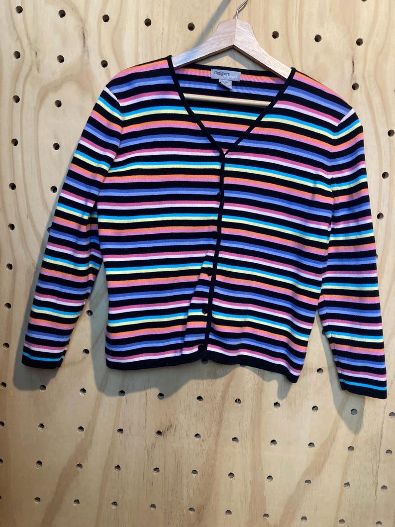 May include: A colourful striped cardigan with a V-neck and long sleeves. The sweater has horizontal stripes in black, pink, blue, yellow, orange, and white. The cardigan is buttoned up and hanging on a wooden hanger.