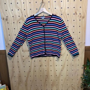 May include: A colourful striped cardigan with a V-neck and long sleeves. The sweater has horizontal stripes in black, blue, pink, orange, and teal. The cardigan is hanging on a wooden hanger.