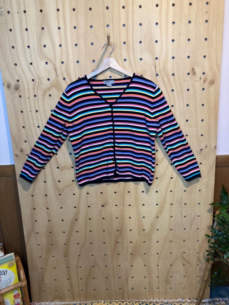 May include: A colourful striped cardigan with a V-neck and long sleeves. The sweater has horizontal stripes in black, blue, pink, orange, and teal. The cardigan is hanging on a wooden hanger.