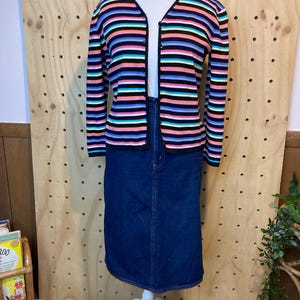 May include: A colourful striped cardigan with a V-neck, worn with a dark blue denim skirt. The cardigan has horizontal stripes in black, pink, orange, blue, and purple. The skirt is knee-length.