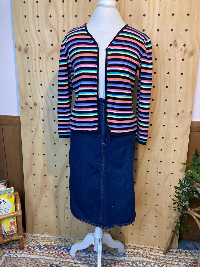 May include: A colourful striped cardigan with a V-neck, worn with a dark blue denim skirt. The cardigan has horizontal stripes in black, pink, orange, blue, and purple. The skirt is knee-length.
