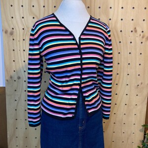 May include: A colourful striped cardigan with a V-neck and long sleeves. The cardigan has horizontal stripes in black, pink, blue, yellow, and orange. The cardigan is buttoned and worn with a denim skirt.