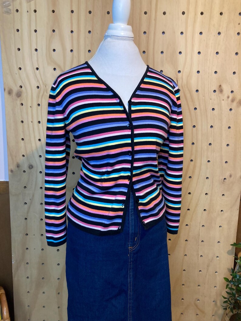 May include: A colourful striped cardigan with a V-neck and long sleeves. The cardigan has horizontal stripes in black, pink, blue, yellow, and orange. The cardigan is buttoned and worn with a denim skirt.