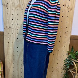 May include: A colourful striped cardigan with a V-neck and button fastening, worn with a dark denim skirt. The cardigan has horizontal stripes in black, pink, blue, yellow, and orange. The skirt is a knee-length, classic denim style.