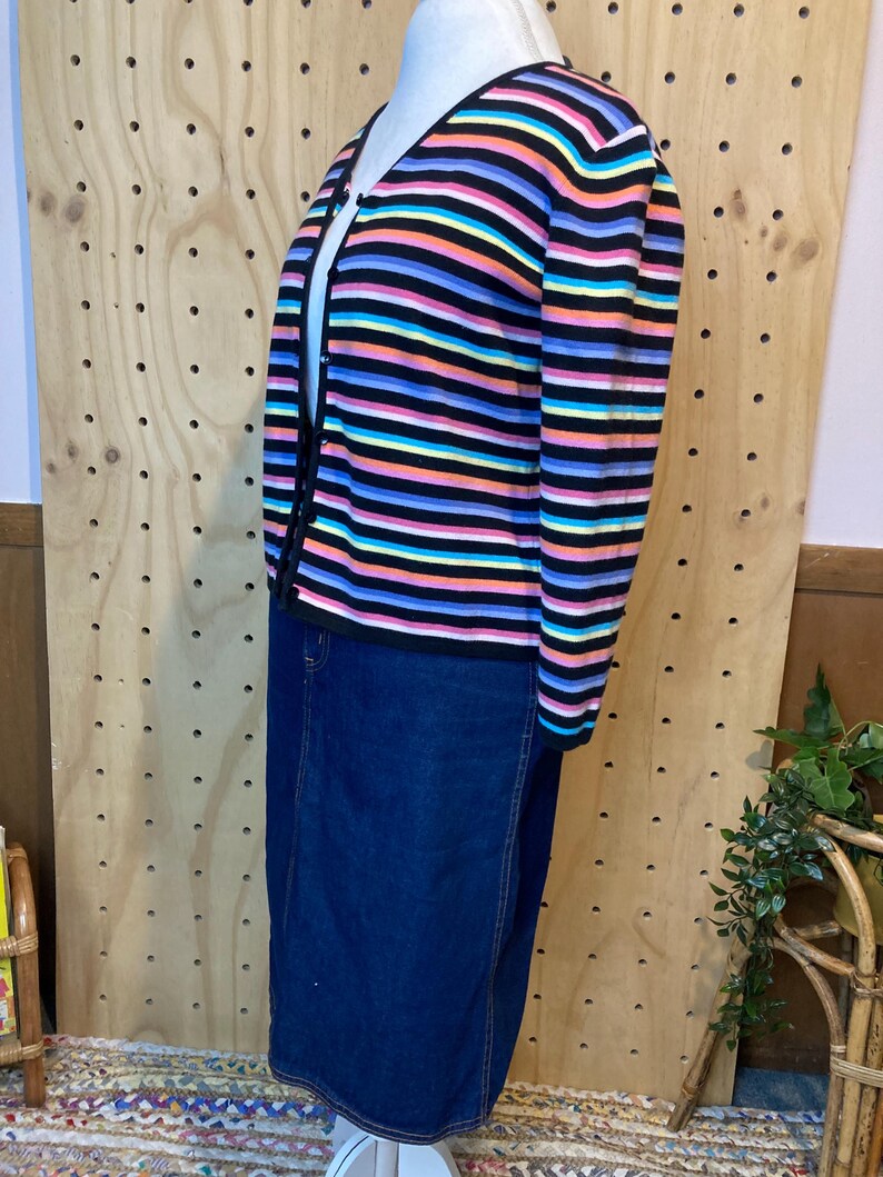 May include: A colourful striped cardigan with a V-neck and button fastening, worn with a dark denim skirt. The cardigan has horizontal stripes in black, pink, blue, yellow, and orange. The skirt is a knee-length, classic denim style.