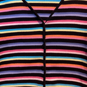 May include: A black cardigan with horizontal stripes in a variety of colours, including pink, orange, yellow, blue, and purple. The sweater has a V-neck and buttons down the front. The overall style is casual and colourful.