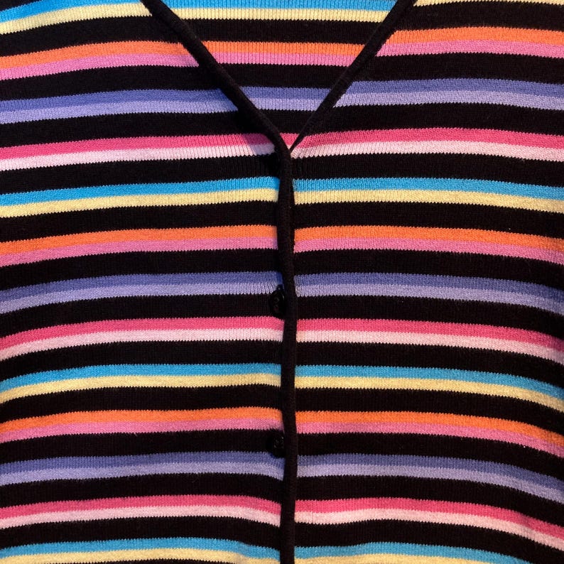 May include: A black cardigan with horizontal stripes in a variety of colours, including pink, orange, yellow, blue, and purple. The sweater has a V-neck and buttons down the front. The overall style is casual and colourful.