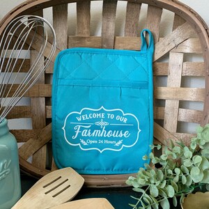 May include: Teal quilted oven mitt with a white design that says "Welcome to our Farmhouse Open 24 Hours".
