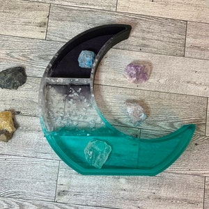 May include: A crescent moon-shaped resin tray with compartments for holding crystals. The tray is teal, black, and white with silver glitter. There are several crystals in the tray and around it, including amethyst, fluorite, and clear quartz.