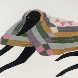 K&ouml;nnte beinhalten: Ein schwarzer Hund mit gro&szlig;en Augen ist in eine bunte Patchwork-Decke gewickelt. Die Decke hat rosa, gr&uuml;ne, gelbe und graue Quadrate mit wei&szlig;en N&auml;hten.