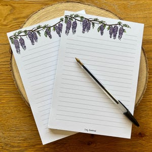 May include: Two lined notepads with a wisteria design and a black pen on a wooden surface. The wisteria illustration features purple flowers and green leaves. The notepads are white with gray lines.