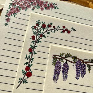 以下が含まれることがあります： 罫線入りの紙と花のコーナーデザインが特徴の文房具セット。デザインには、ピンクの桜、赤いバラ、紫の藤が含まれています。紙はオフホワイトで、黒い横線が入っています。