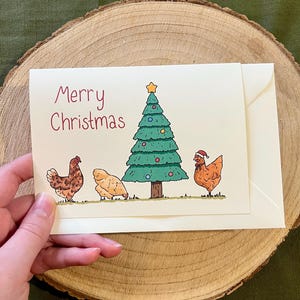 Puede incluir: Tarjeta navideña crema con la frase "Merry Christmas" en escritura roja. La tarjeta presenta una ilustración de dibujos animados de un árbol de Navidad y tres gallinas. Una gallina lleva un gorro de Papá Noel.
