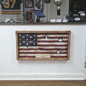 Könnte beinhalten: Gerahmte amerikanische Flagge aus Holz mit einem Used-Look, mit roten und weißen Streifen und einem blauen Feld mit weißen Sternen. Die Flagge hat mehrere Einschusslöcher und eine kleine Tafel mit Text. Ein rustikales, patriotisches Wohnaccessoire.