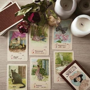 Può includere: Un mazzo di carte dei tarocchi con illustrazioni ad acquerello, tra cui un gatto nero, una donna in un giardino e una farfalla. Il mazzo è accompagnato da rose secche, candele e un cofanetto con il testo "I misteri della Sibilla".