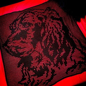 Può includere: Una coperta all'uncinetto nera e rossa con un design pixelato. La coperta presenta un grande disegno centrale nero su uno sfondo rosso. La coperta è di forma quadrata e sembra fatta a mano.
