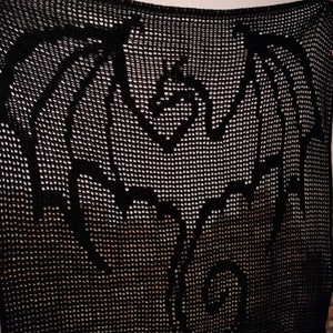 Può includere: Coperta nera all'uncinetto con un motivo a drago. Il drago è raffigurato con le ali spiegate e la coda arricciata. La coperta è realizzata con filato scuro, possibilmente nero, e il disegno è creato dall'intreccio aperto dell'uncinetto.