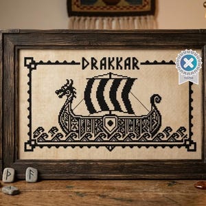 Può includere: Un ricamo a punto croce incorniciato raffigurante una nave vichinga nera su uno sfondo di tessuto beige. La parola "DRAKKAR" è ricamata in nero sopra la nave, circondata da un bordo geometrico. Un emblema circolare blu si trova in alto a destra.