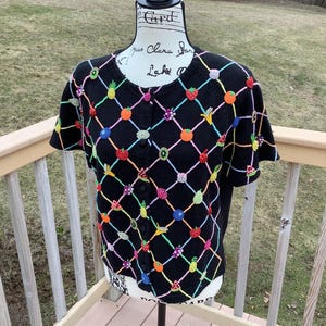 May include: Black short-sleeved cardigan with a colorful beaded fruit design. The cardigan features a grid pattern with various fruits like strawberries, oranges, and bananas. It has a button-up front and is displayed on a mannequin.