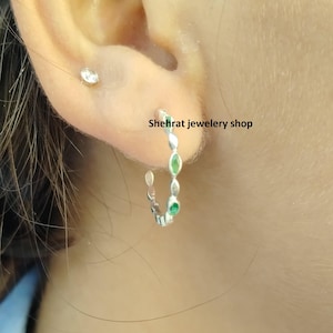 Può includere: Orecchini a cerchio in argento con piccole pietre preziose verdi. Gli orecchini sono mostrati sull'orecchio di una persona.